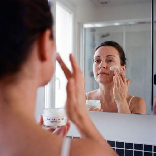 Crème visage au lait de chèvre appliquée sur le visage d’une femme à la peau mature devant un miroir