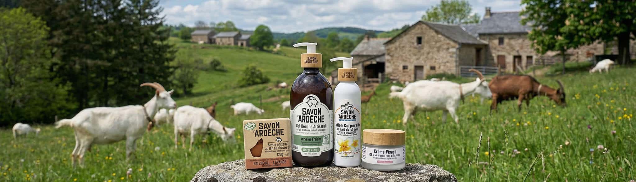 Produits Savon d’Ardèche au lait de chèvre posés dans une prairie ardéchoise, avec des chèvres en arrière-plan